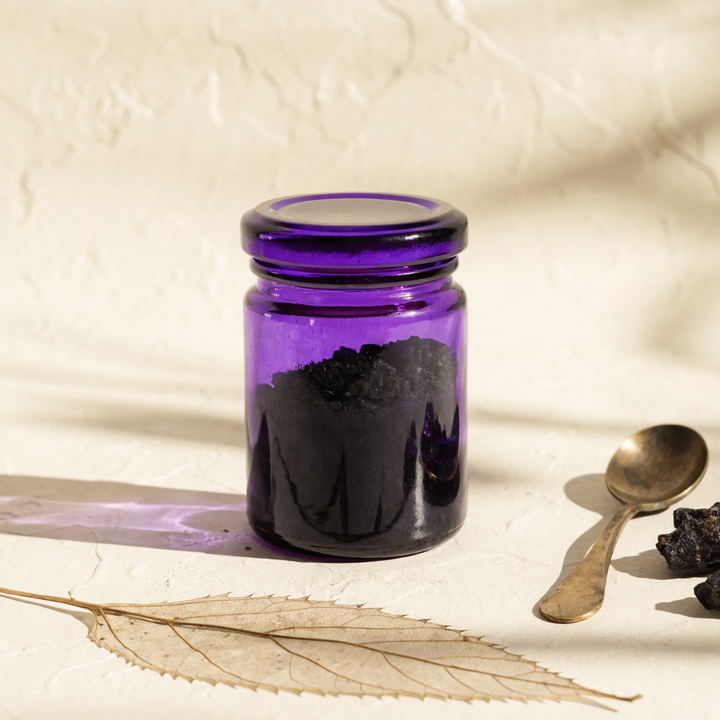 Apothecary still-life: Miron violet glass jar of pure Altai shilajit resin with antique brass spoon, mineral fragments, and dried botanical accent on cream plaster surface